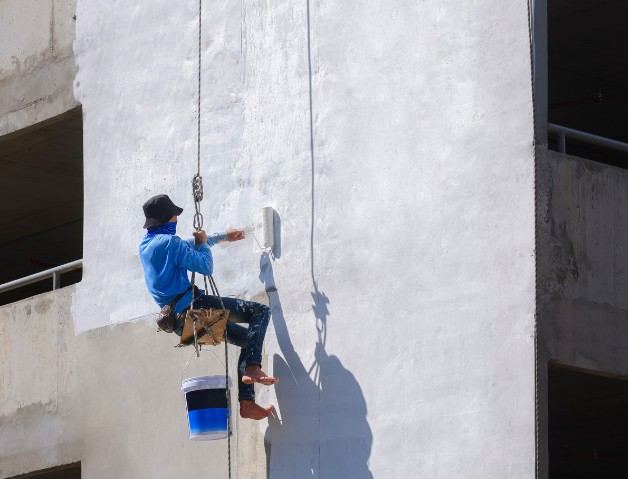Pintado sin andamio en Mallorca Operario colgado de una cuerda pintando con rodillo una fachada blanca: trabajos verticales de pintura